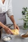 Panadero femenino anónimo en delantal amasando masa blanda con harina en la mesa cerca de la salsa de manzana y ramo de flores mientras cocina pastelería - foto de stock