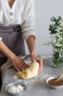 Anonimo panettiere femminile in grembiule impastando pasta morbida con farina sul tavolo vicino alla salsa di mele e bouquet di fiori durante la cottura della pasta — Foto stock
