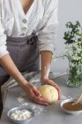 Boulangère anonyme en tablier pétrissant pâte molle avec farine sur la table près de la compote de pommes et bouquet de fleurs pendant la cuisson de la pâtisserie — Photo de stock