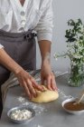 Anonymous female baker in apron kneading soft dough with flour on table near apple sauce and bouquet of flowers while cooking pastry — Stock Photo