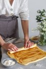 Mulher anônima padeiro em avental fazendo rolo de massa macia com molho de maçã na mesa perto de farinha e buquê de flores — Fotografia de Stock