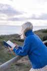 Side view of unrecognizable young female in blue sweater leaning on wooden fence and enjoying favorite poetry while spending time at seaside — Stock Photo