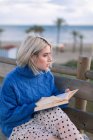Side view of young blonde female in warm blue sweater and skirt looking away while sitting on wooden bench in terrace against blurred beach and reading book — Stock Photo