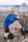 Junge Frau in blauem Pullover und Rock sitzt auf Holzbank und streichelt Hund, während sie mit Buch am Meer ausruht und wegschaut — Stockfoto