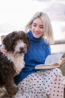 Junge Frau in blauem Pullover und Rock sitzt auf Holzbank und streichelt Hund, während sie sich mit Buch am Meer ausruht — Stockfoto
