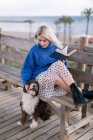 Joven hembra en jersey azul y falda sentada en banco de madera y perro acariciando mientras descansa con libro en la playa - foto de stock