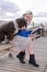 Junge Frau in blauem Pullover und Rock sitzt mit Hund auf Holzbank, während sie mit Buch am Meer ruht — Stockfoto