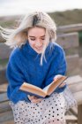 Young blonde female in warm blue sweater and skirt looking away while sitting on wooden bench in terrace against blurred beach and reading book — Stock Photo