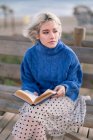 Молода блондинка в теплий блакитний светр і спідниця, дивлячись далеко, сидячи на дерев'яній лавці в терасі проти розмитого пляжу і читання книги — стокове фото