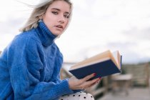 Вид сбоку молодой блондинки в теплом синем свитере и юбке, смотрящей в камеру, сидя на деревянной скамейке на террасе на размытом фоне книги для чтения — стоковое фото