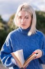 Задумчивая молодая блондинка в тёплом голубом свитере, опирающаяся на деревянный забор и читающая книгу, проводя весенний день в сельской местности, отворачиваясь — стоковое фото