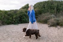 Side view of young blonde female in casual sweater and skirt with book in hand walking with dog on sandy beach near green plants — Stock Photo