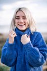 Gioioso allegro giovane donna bionda in maglione blu lavorato a maglia guardando la fotocamera e ridendo mentre in piedi contro lo sfondo sfocato della costa del mare — Foto stock