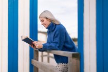 Happy young female in warm sweater and skirt leaning on wooden fence with open book reading against white and blue striped wall — Stock Photo