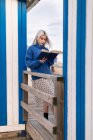 Nachdenkliche junge Frau in warmem Pullover und Rock lehnt an Holzzaun mit aufgeschlagenem Buch lesen an weiß-blau gestreifter Wand — Stockfoto