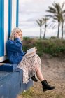 Vista lateral da jovem mulher pensativa em suéter quente e saia sentada com livro aberto lendo contra a parede listrada branca e azul olhando para longe — Fotografia de Stock