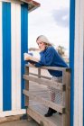 Giovane donna premurosa in maglione caldo e gonna appoggiata sulla recinzione in legno con lettura a libro aperto contro la parete a strisce bianche e blu — Foto stock