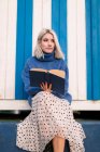 Знизу вдумлива молода жінка в теплому светрі і спідниці сидить з відкритою книгою читання проти білої і синьої смугастої стіни, дивлячись далеко — стокове фото
