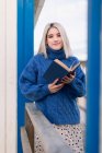 Mujer joven pensativa en suéter cálido y falda sonriendo y mirando a la cámara mientras se apoya en una valla de madera con libro abierto contra la pared de rayas blancas y azules - foto de stock