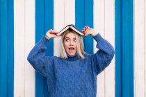 Смешно удивленная молодая женщина в обычном трикотажном свитере держит открытую книгу на голове и смотрит в сторону, стоя напротив красочной стены — стоковое фото