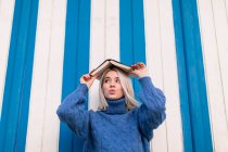 Смешно удивленная молодая женщина в обычном трикотажном свитере держит открытую книгу на голове и смотрит в сторону, стоя напротив красочной стены — стоковое фото