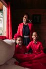 Cheerful young black female with cute laughing siblings in red clothes looking at camera while spending time together in cozy country house — Stock Photo
