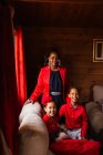 Joyeux jeune femme noire avec des frères et sœurs riants mignons en vêtements rouges regardant la caméra tout en passant du temps ensemble dans une maison de campagne confortable — Photo de stock