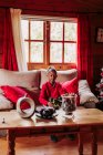 Удовлетворенная черная девушка держит игрушку в руках и смотрит в камеру, сидя на диване возле окна в уютной гостиной с рождественским декором — стоковое фото