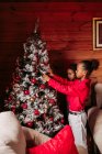 Side view of cute ethnic siblings in casual clothes decorating traditional Christmas tree in cozy wooden house — Stock Photo