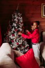Vista lateral de irmãos étnicos bonitos em roupas casuais decorando a árvore de Natal tradicional na acolhedora casa de madeira — Fotografia de Stock