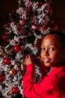 Vista laterale di allegra bambina nera in camicia rossa guardando la fotocamera e sorridendo mentre in piedi vicino all'albero di Natale decorato a casa — Foto stock