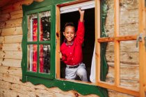 Criança preta alegre em camisa vermelha e calças brancas olhando para a câmera através da janela aberta da casa de madeira rural — Fotografia de Stock