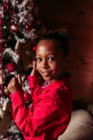 Вид збоку весела маленька чорна дівчинка в червоній сорочці дивиться на камеру і посміхається, стоячи біля прикрашеної ялинки вдома — стокове фото