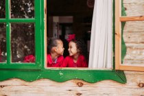 Niedliche schwarze kleine Geschwister lächeln durch das offene Fenster der Holzhütte — Stockfoto