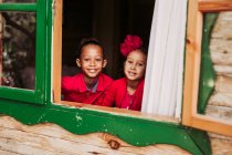 Joli petit frère noir souriant et regardant la caméra à travers la fenêtre ouverte de la cabine en bois — Photo de stock