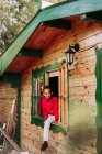Веселый черный ребенок в красной рубашке и белых брюках смотрит в камеру через открытое окно деревенского деревянного дома — стоковое фото