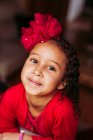 From above of cute little curly haired ethnic girl with red bow wearing red dress looking at camera and smiling against blurred background — Stock Photo