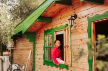 Cheerful black child in red shirt and white pants looking through open window of rural wooden house — Stock Photo