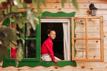 Fröhliches schwarzes Kind in rotem Hemd und weißer Hose blickt durch das offene Fenster eines ländlichen Holzhauses — Stockfoto
