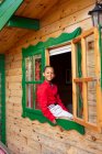Cheerful black child in red shirt and white pants looking at camera through open window of rural wooden house — Stock Photo