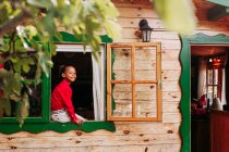 Cheerful black child in red shirt and white pants looking through open window of rural wooden house — Stock Photo