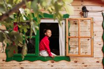 Criança preta alegre em camisa vermelha e calças brancas olhando através da janela aberta da casa de madeira rural — Fotografia de Stock