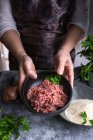 Femme méconnaissable dans un tablier démontrant bol avec de la viande fraîche hachée et persil à la caméra lors de la cuisson sur table grise — Photo de stock