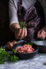 Обрезанная неузнаваемая женщина в фартуке держит кучу свежих листьев над миской с сырым мясом во время приготовления обеда на кухне — стоковое фото