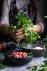Unerkennbare Frau mit Schürze hält Bündel frischer Petersilie über Schüssel mit rohem Hackfleisch, während sie das Mittagessen in der Küche kocht — Stockfoto