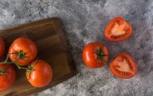Halved and whole fresh tomatoes placed wooden board on rough gray table during food preparation in kitchen — Stock Photo