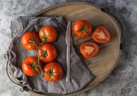 Tomates frescos cortados pela metade e inteiros colocados tábua de madeira em mesa cinza áspera durante a preparação de alimentos na cozinha — Fotografia de Stock