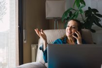 Mujer adulta con portátil sonriendo y gesticulando mientras mantiene una conversación en el teléfono inteligente durante el trabajo en casa - foto de stock