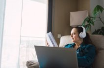 Mature woman listening to music in headphones and reading paper while sitting in armchair and doing remote job on laptop near window at home — Stock Photo