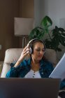 Mujer madura escuchando música en auriculares y leyendo papel mientras está sentada en un sillón y haciendo un trabajo remoto en la computadora portátil cerca de la ventana en casa - foto de stock